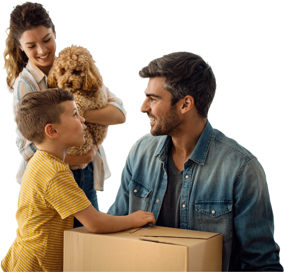 A woman holding a small dog stands next to a smiling man and a young boy, who are near a large cardboard box.