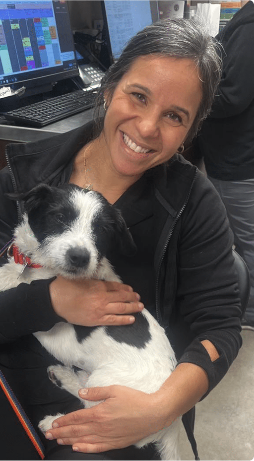 A woman sitting indoors smiles while holding a small black and white dog in her arms; computer screens are visible in the background.