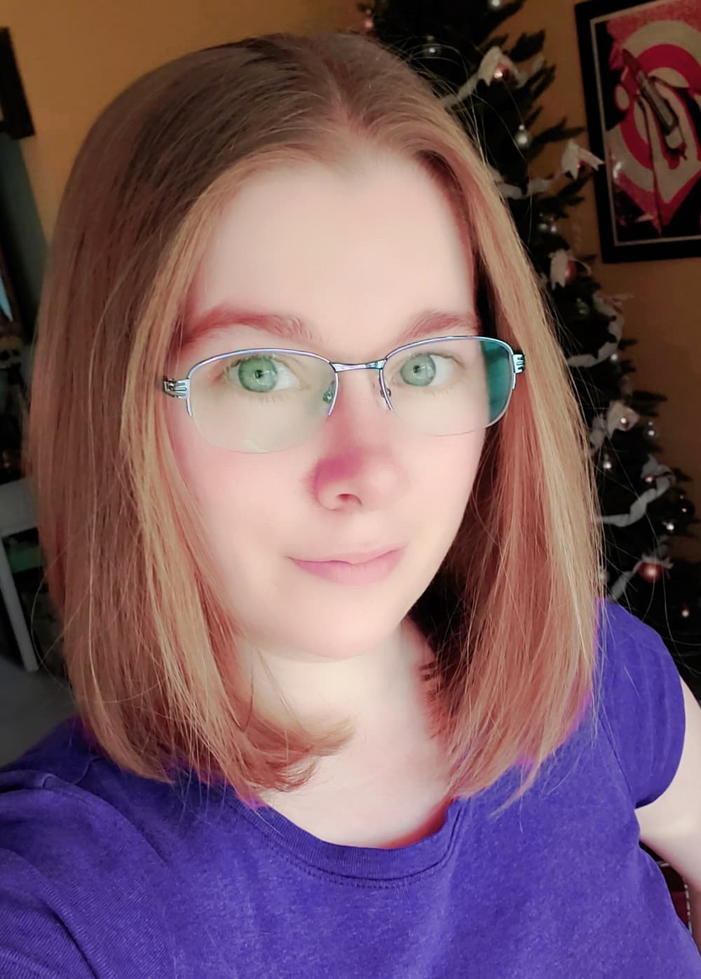 Person with straight, shoulder-length light brown hair and glasses, wearing a purple shirt, stands indoors with a decorated Christmas tree in the background.