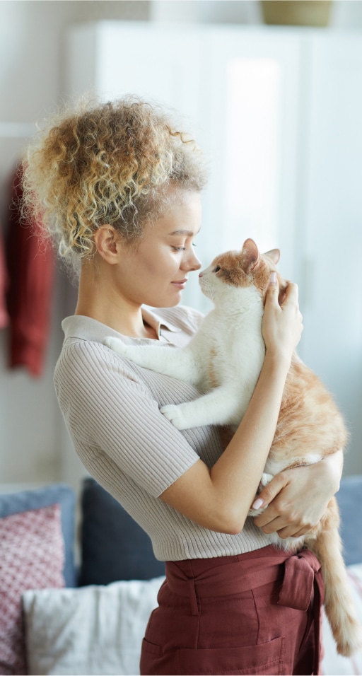 A woman with curly hair holds an orange and white cat, looking at it closely in a well-lit indoor setting.