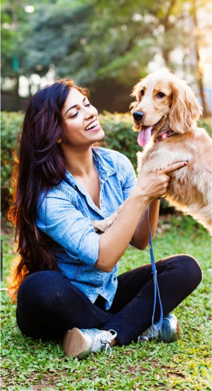 A woman sitting on grass outdoors holds a golden retriever puppy on her lap, both looking happy.